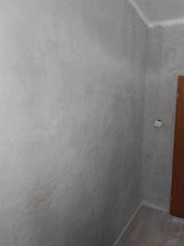 Szara ściana po nałożeniu gładzi szpachlowej, widoczna faktura i drobne nierówności, przygotowana do malowania, fragment drewnianej podłogi i drzwi w tle.