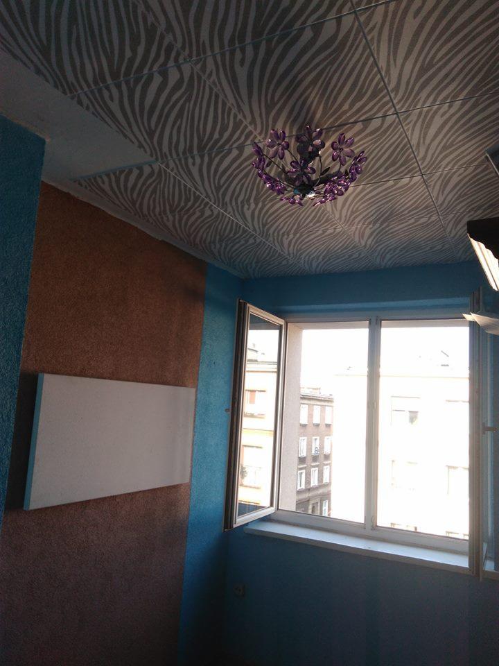 Pokój z dekoracyjnym sufitem w zebrę i fioletowym żyrandolem, niebieskimi ścianami i otwartym oknem z widokiem na budynki miejskie.