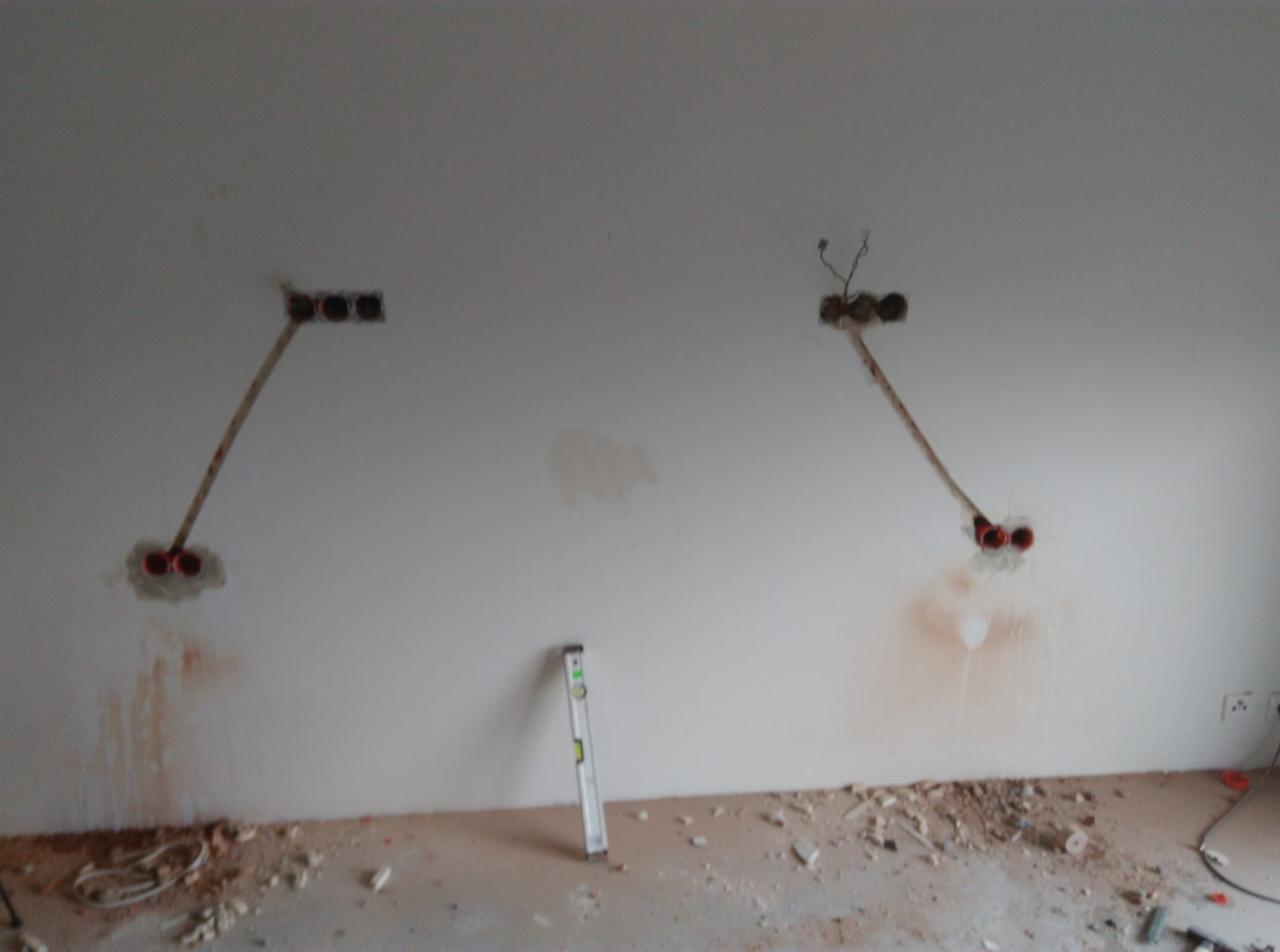 Ściana w trakcie remontu z widocznymi dwoma miejscami po kuciu pod instalację elektryczną, wystające kable i rurki, poziomica oparta o ścianę, gruz na podłodze.