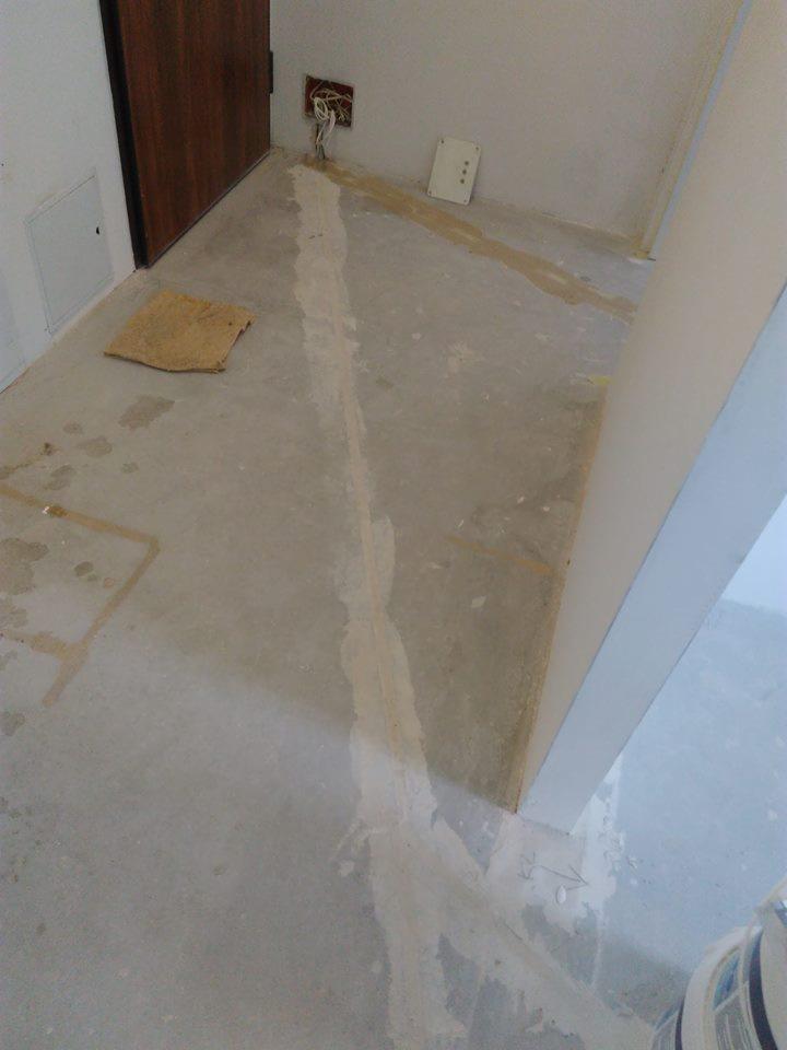 Przygotowanie podłoża pod posadzkę: wylany grunt wyrównujący na betonowej powierzchni, widoczne ślady po rozprowadzaniu, otwarta puszka elektryczna z kablami w tle.