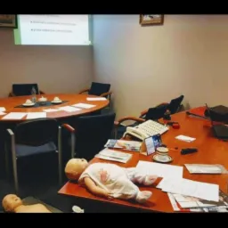 Sala konferencyjna z owalnym stołem, rzutnikiem wyświetlającym prezentację, manekin niemowlęcia leżący na biurku, prawdopodobnie podczas kursu pierwszej pomocy.