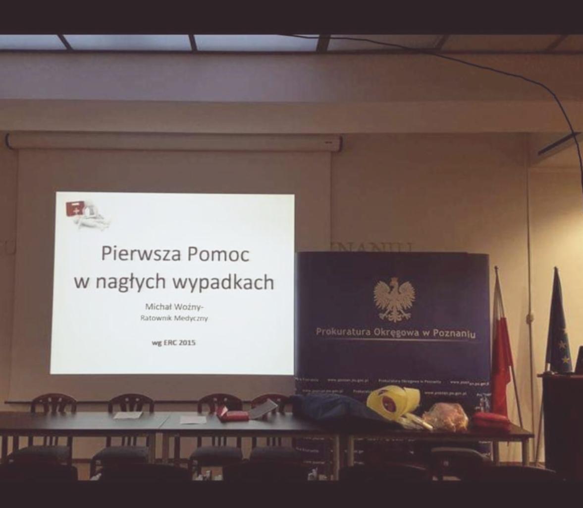 Sala konferencyjna z rzutnikiem wyświetlającym slajd prezentacji 'Pierwsza Pomoc w nagłych wypadkach', obok flaga Polski i flaga Unii Europejskiej oraz baner 'Prokuratura Okręgowa w Poznaniu'