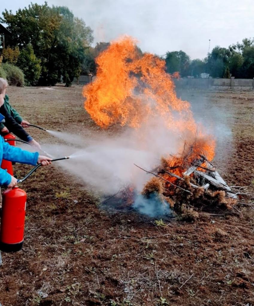 Osoby w trakcie gaszenia ogniska z gałęzi przy użyciu gaśnic podczas szkolenia przeciwpożarowego na zewnątrz.