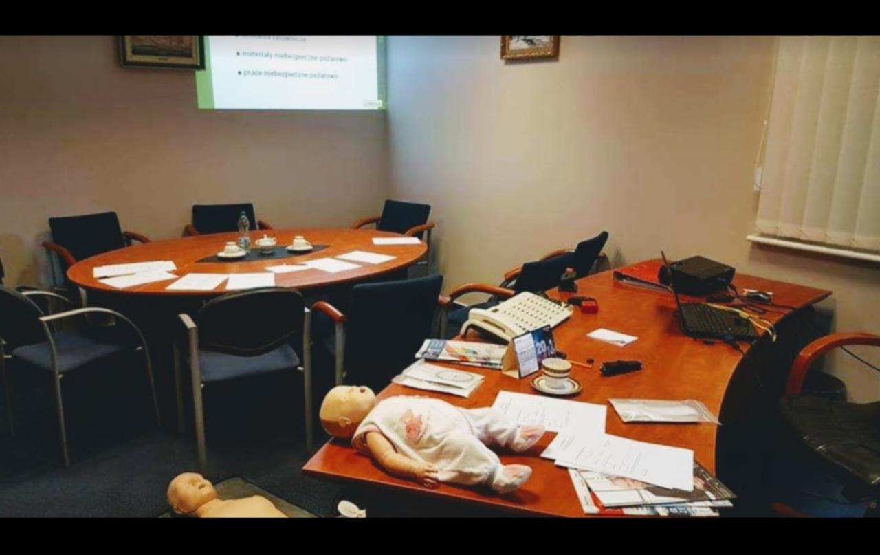 Sala konferencyjna z owalnym stołem, rzutnikiem wyświetlającym prezentację, manekin niemowlęcia leżący na biurku, prawdopodobnie podczas kursu pierwszej pomocy.