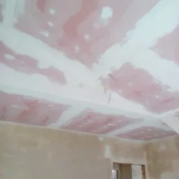 Nieukończone łączenia płyt kartonowo-gipsowych na suficie, widoczne ślady szpachli w kolorze różowym, surowe ściany przygotowane do dalszych prac wykończeniowych, fragment otworu drzwiowego.