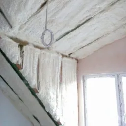 Poddasze z widoczną izolacją natryskową z pianki poliuretanowej, eksponowane krokwie dachowe, białe okno PCV w tle, przewód elektryczny zwisający z sufitu.