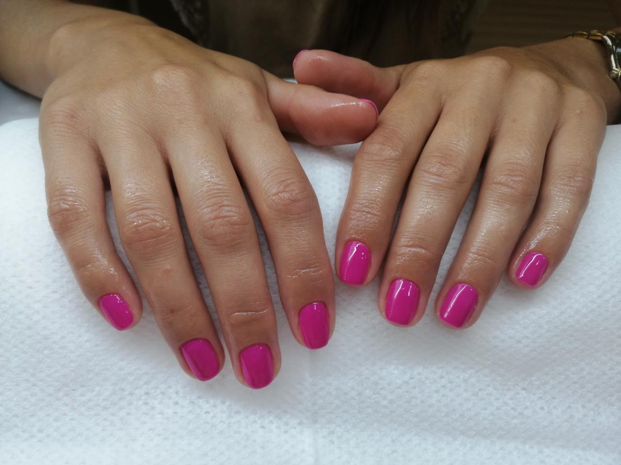 Dłonie kobiety z świeżo pomalowanymi paznokciami w kolorze intensywnego różu na białym ręczniku. Widoczne delikatne krople oliwki na skórze dłoni.