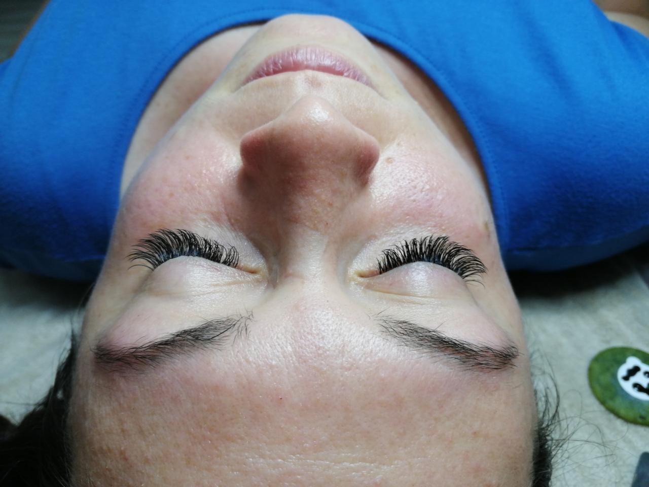 Zbliżenie na twarz kobiety z zamkniętymi oczami po zabiegu przedłużania rzęs; widoczne długie, gęste, czarne rzęsy, naturalny makijaż brwi, niebieska koszulka i zielony kamień w tle.