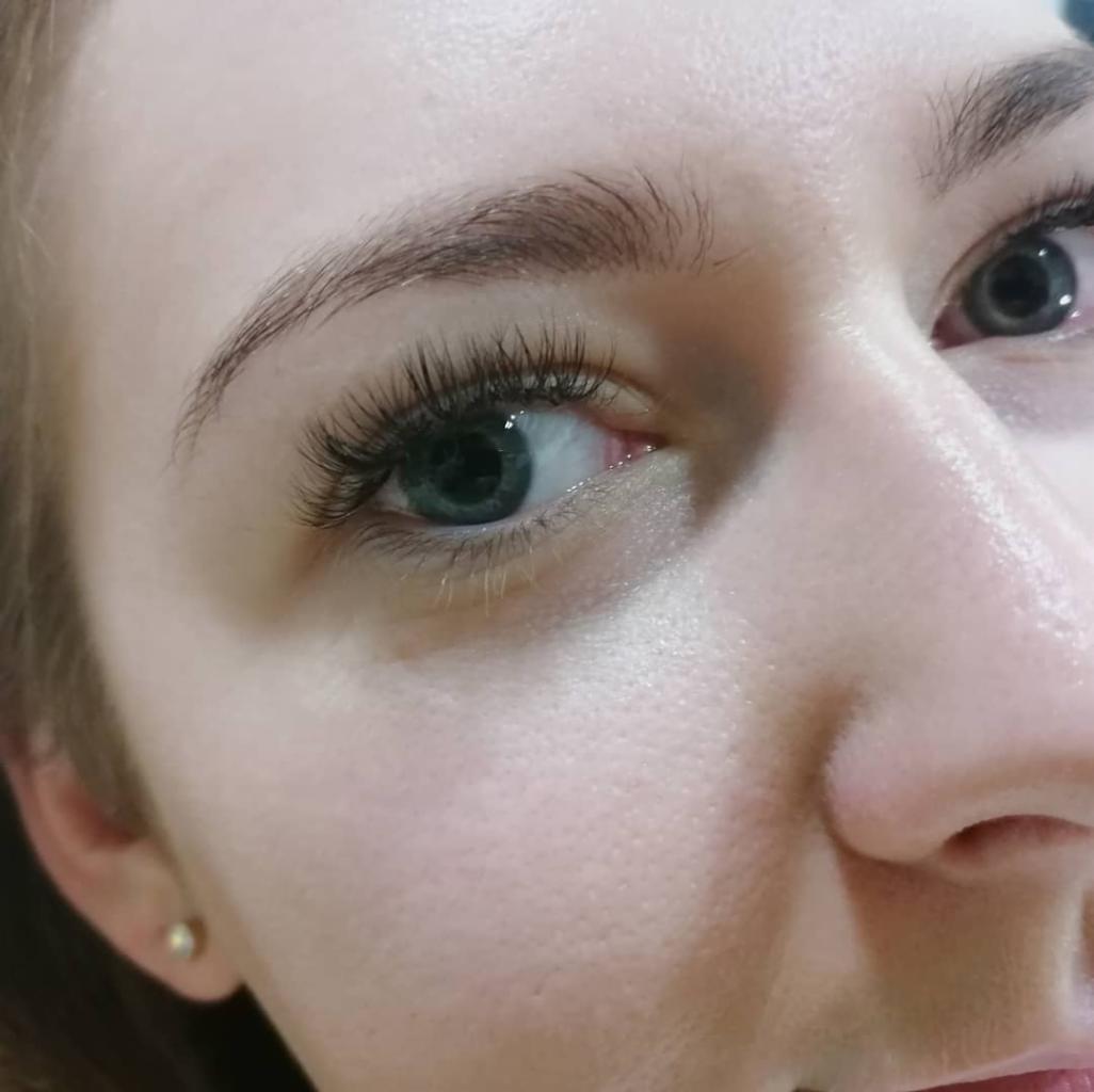 Zbliżenie na kobiece oko z wyraźnie przedłużonymi rzęsami, widoczny delikatny makijaż brwi i złota kolczyk w uchu.