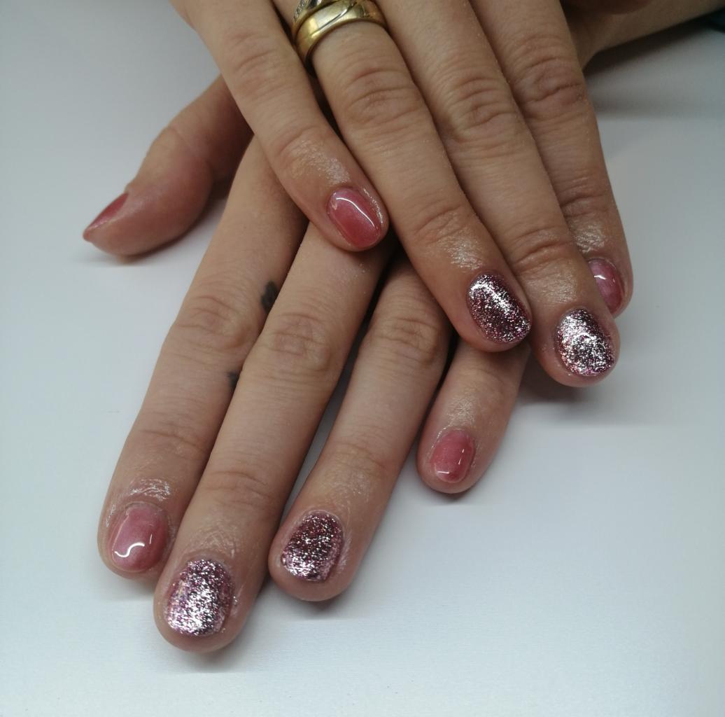 Dłonie kobiety z manicure hybrydowym w odcieniach różu i brokatu, wykonanym w Rzeszowie, na białym tle.