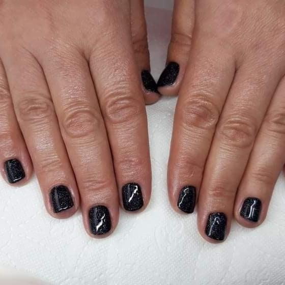 Dłonie z krótkimi paznokciami pomalowanymi ciemnym, brokatowym lakierem; na paznokciach widoczne delikatne, białe refleksy świetlne.