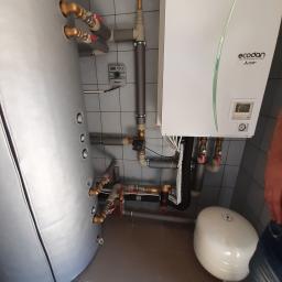 ekoRyniec s.c. M.D. Ryniec - Instalacja pompy ciepła Ecodan z szarym zbiornikiem akumulacyjnym, białym zbiornikiem przeponowym i szarą instalacją hydrauliczną w pomieszczeniu z szarymi płytkami.