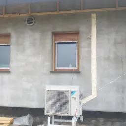 Zewnętrzna jednostka klimatyzacji Daikin zamontowana na białym stelażu przy szarej ścianie budynku w trakcie prac wykończeniowych, widoczne rury w izolacji z pianki montażowej, okna z brązowymi...