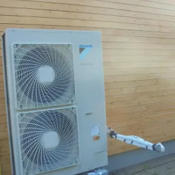 Zewnętrzna jednostka pompy ciepła Daikin Altherma zamontowana na betonowych bloczkach przed drewnianą ścianą budynku, widoczne rury instalacyjne.