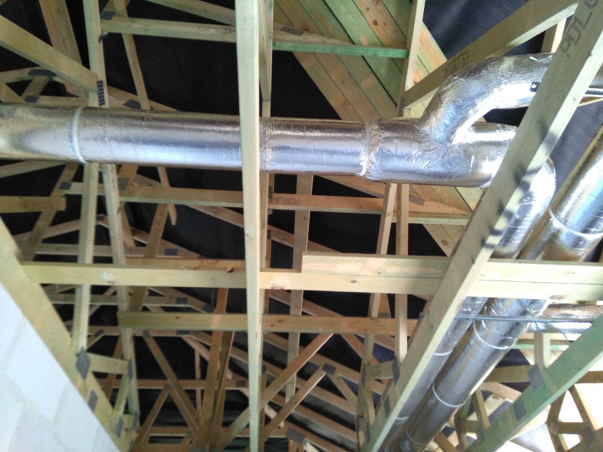 Instalacja przewodów wentylacyjnych w izolacji aluminiowej, widok z dołu na drewnianą konstrukcję dachu i czarną membranę dachową.