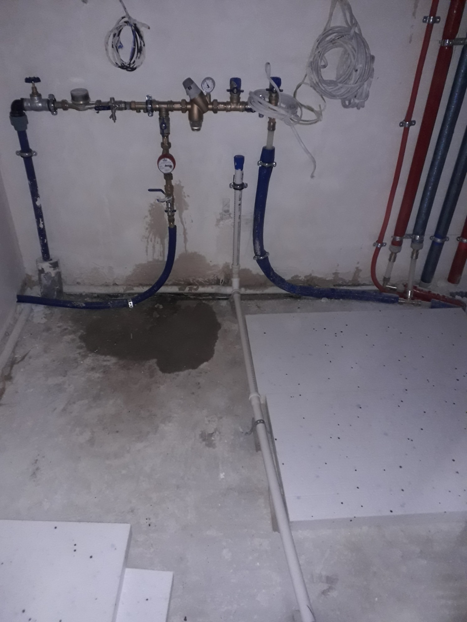 Surowy montaż instalacji wodnej z manometrami, zaworami i niebieskimi rurami, widoczne fragmenty izolacji podłogowej i ślady wilgoci na podłodze.
