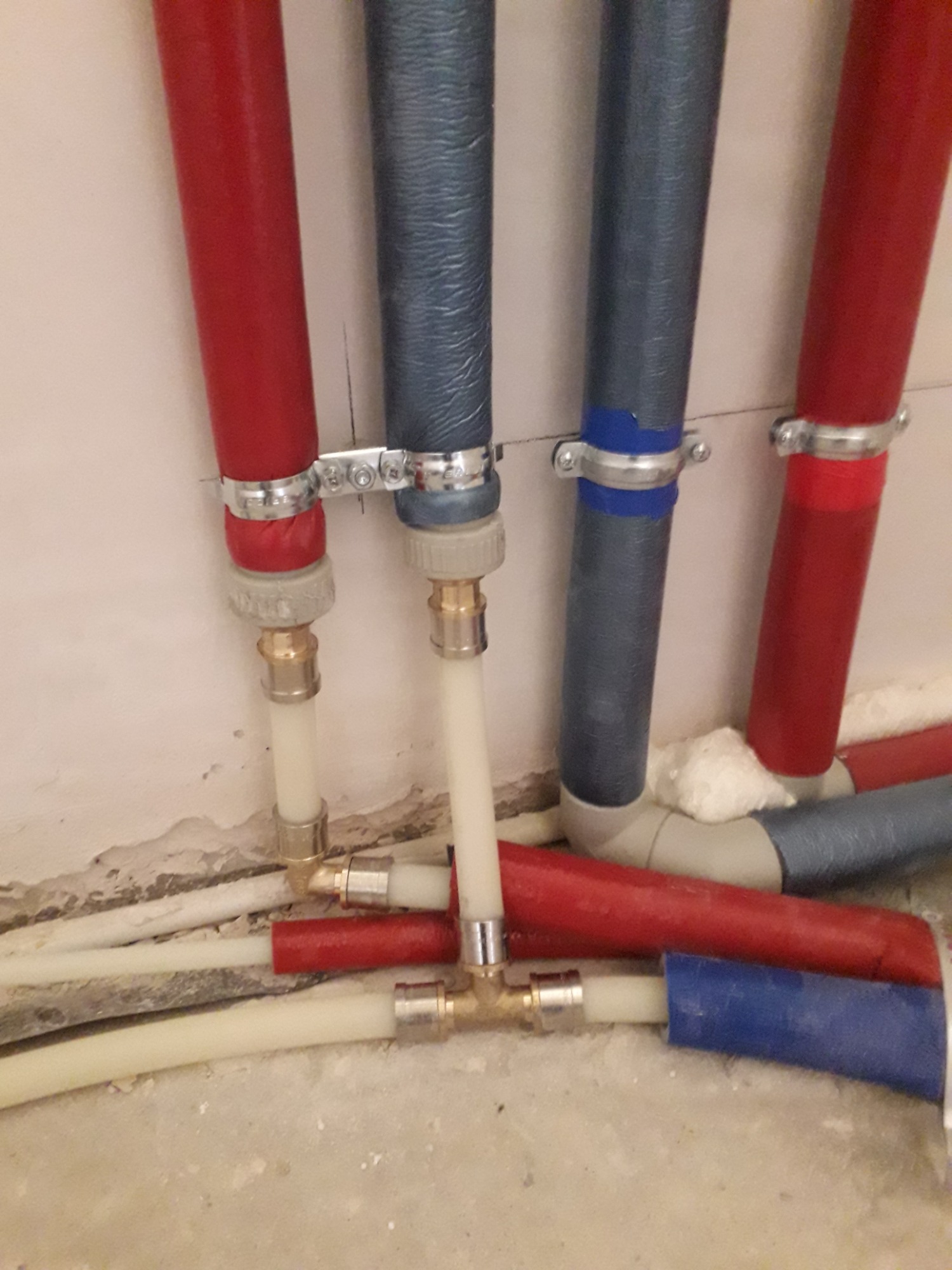 Połączenie rur instalacyjnych PEX w kolorze czerwonym, niebieskim i białym, zabezpieczonych metalowymi obejmami, widoczne złącza mosiężne i izolacja piankowa, ułożone wzdłuż ściany.