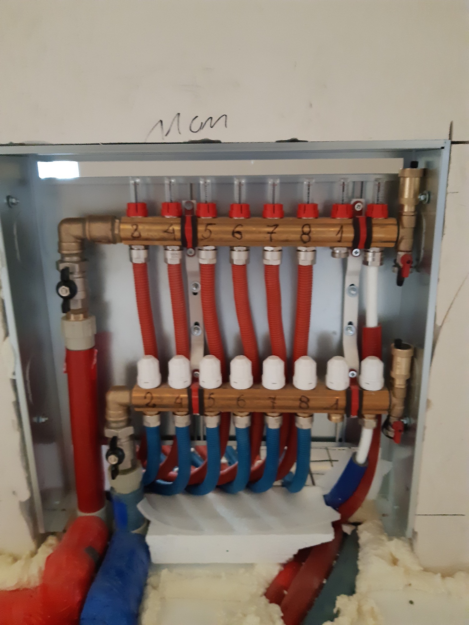 Rozdzielacz ogrzewania podłogowego w metalowej szafce, z widocznymi rurami w kolorach czerwonym i niebieskim, ponumerowanymi zaworami i izolacją z pianki montażowej.