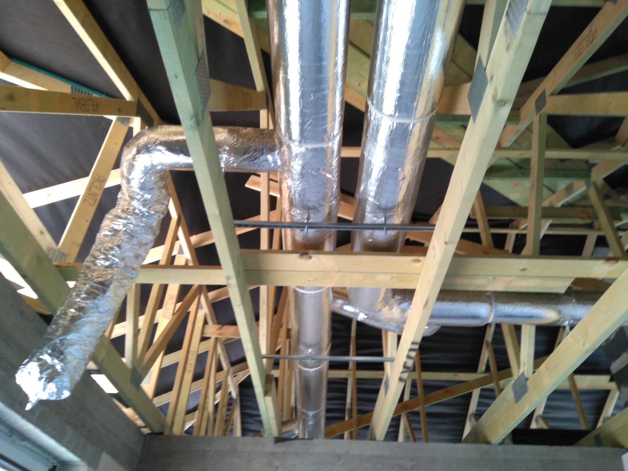 Srebrne, izolowane kanały wentylacyjne zamontowane w drewnianej konstrukcji dachu z czarną membraną dachową w tle.