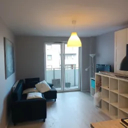 Wnętrze pokoju po remoncie: ściany pomalowane na szaro, jasna podłoga, widoczny fragment sofy, regał i lampa wisząca.