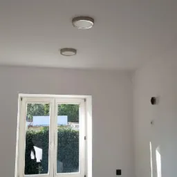 Wnętrze pokoju w trakcie prac wykończeniowych, widoczne trzy okrągłe lampy sufitowe, okno z widokiem na zieleń, ściany pomalowane na biało, widoczne gniazdka elektryczne.