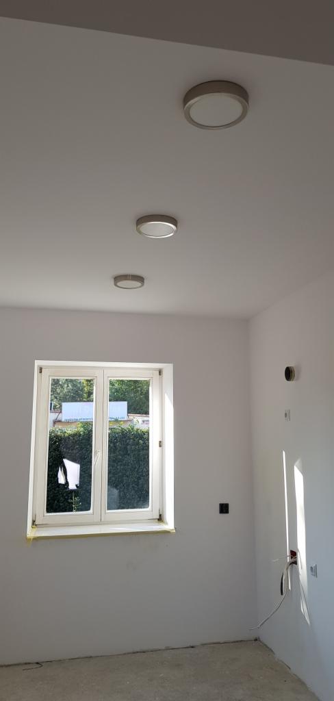 Wnętrze pokoju w trakcie prac wykończeniowych, widoczne trzy okrągłe lampy sufitowe, okno z widokiem na zieleń, ściany pomalowane na biało, widoczne gniazdka elektryczne.