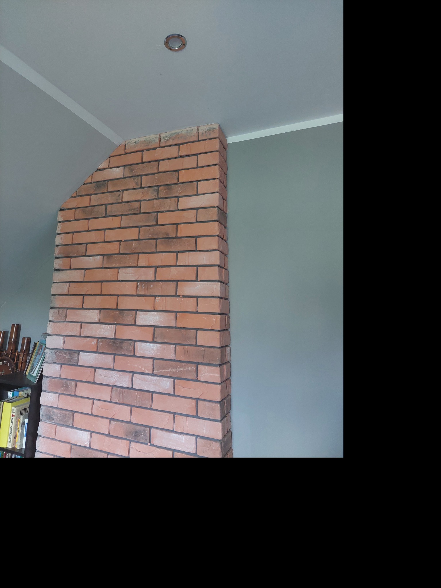 Ściana z cegły klinkierowej w rogu pokoju, pomalowana szara ściana w tle. Widoczne oświetlenie sufitowe i fragment regału z książkami.