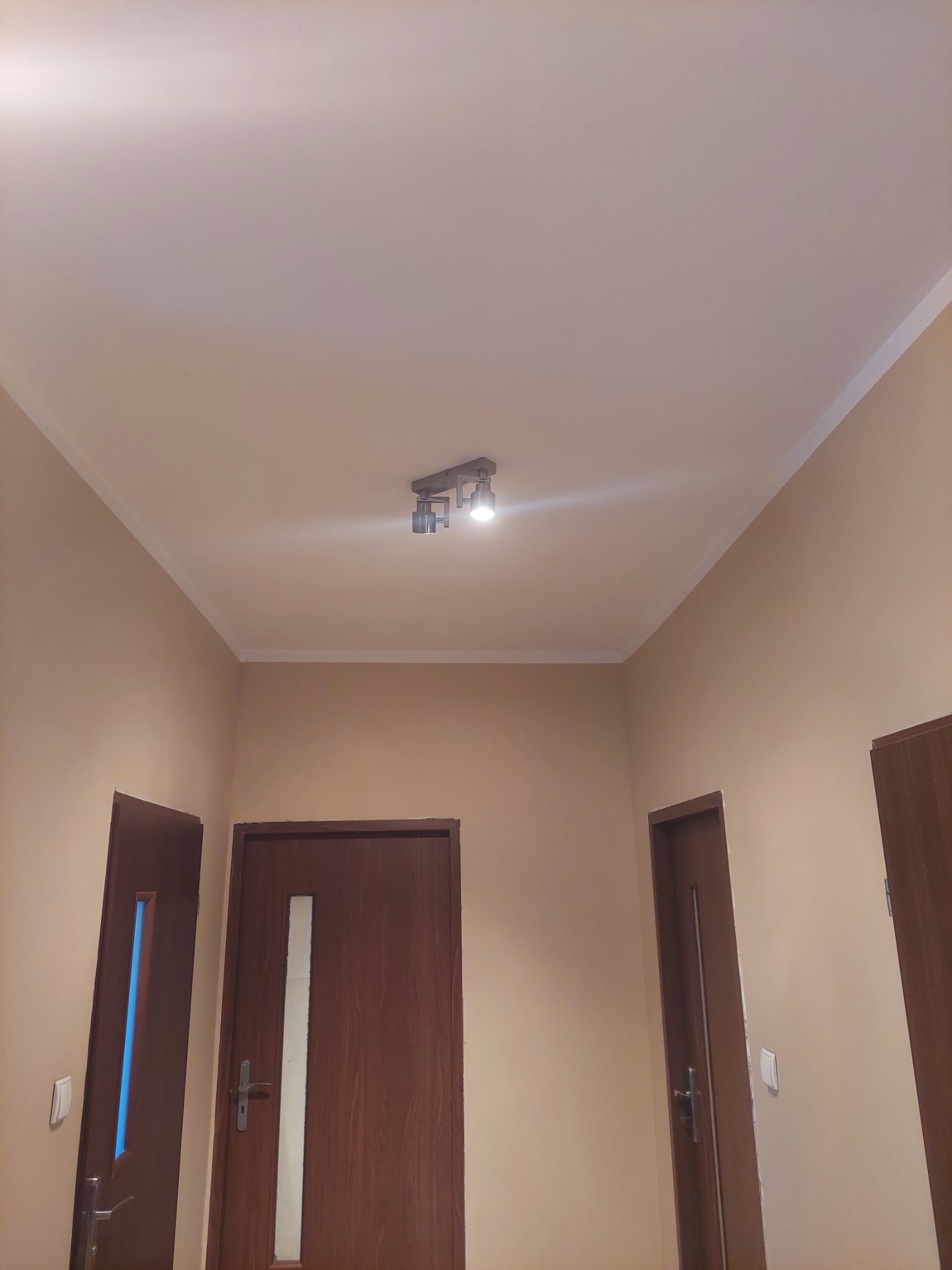 Beżowe ściany i sufit po malowaniu w korytarzu z trzema parami drzwi. Listwa dekoracyjna pod sufitem. Na suficie lampa halogenowa.