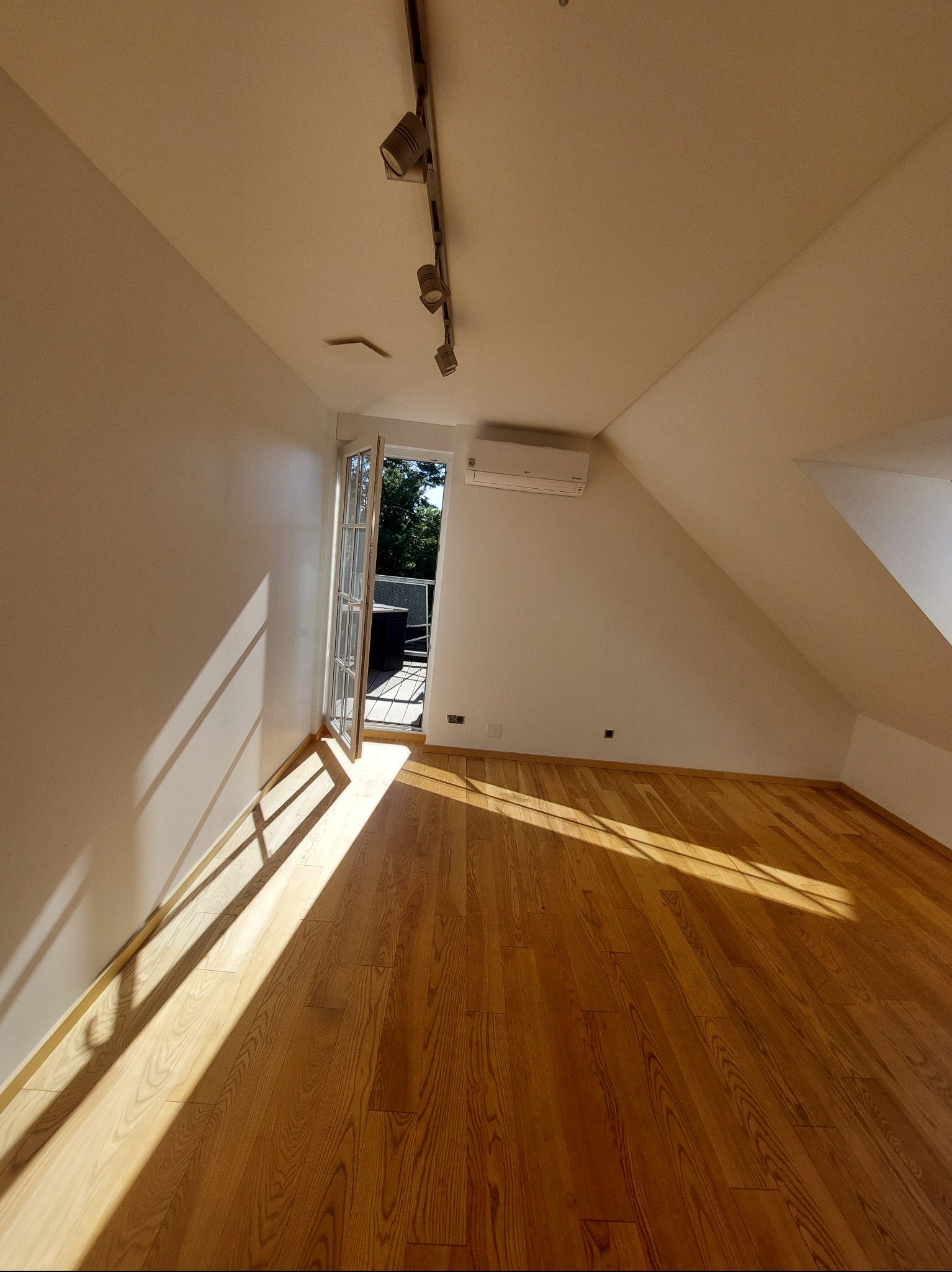 Jasne poddasze po odświeżeniu: białe ściany, drewniana podłoga w ciepłym odcieniu, otwarte drzwi balkonowe wpuszczające promienie słoneczne. Minimalistyczne oświetlenie.