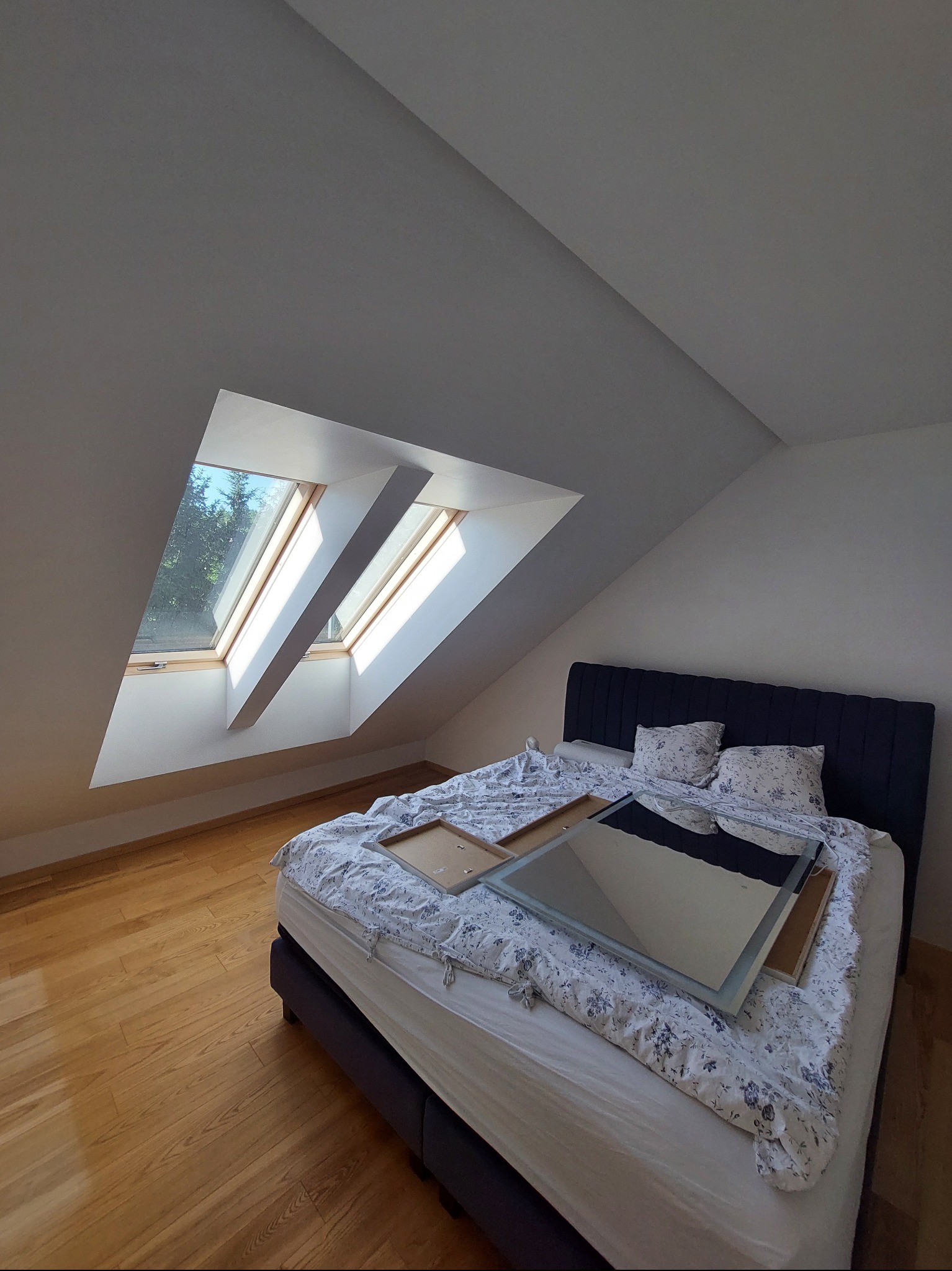Poddasze z łóżkiem przygotowane do malowania: okna dachowe z widokiem na zieleń, zabezpieczone meble, drewniana podłoga. Pokój czeka na odświeżenie koloru.