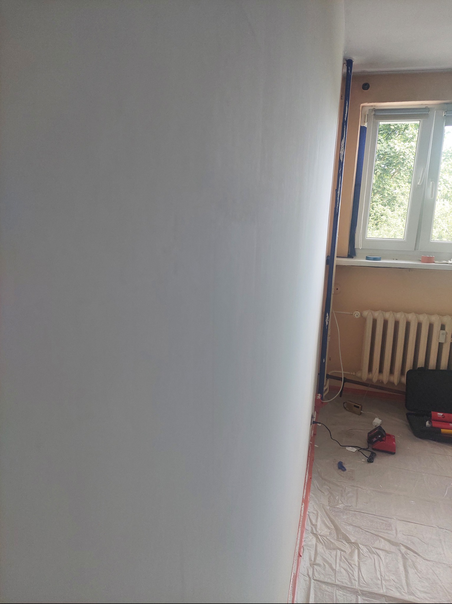 Ściana pomalowana na biało, zabezpieczona folią i taśmą malarską przy oknie. Widoczny grzejnik, okno i narzędzia malarskie. Etap prac wykończeniowych w pokoju.