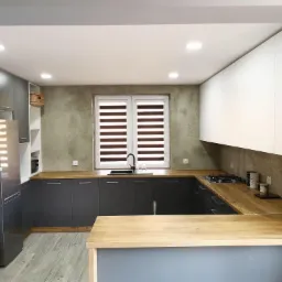 Nowoczesna kuchnia z szarymi szafkami dolnymi, białymi górnymi i drewnianym blatem, widoczne okno z roletami dzień-noc i betonowa ściana w tle.