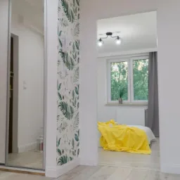 Biała szafa przesuwna z lustrem w przedpokoju, widok na sypialnię z żółtą narzutą i tapetą w liście.