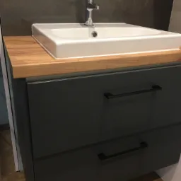 Szara szafka łazienkowa z dwiema szufladami, drewnianym blatem i białą umywalką z chromowaną baterią, na tle szarej ściany.