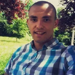 Portret uśmiechniętego mężczyzny w niebiesko-szarej koszuli w kratę na tle zieleni parkowej.