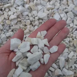 Otwarta dłoń trzymająca w garści biały, drobny kamień ozdobny na tle większej ilości tego samego kruszywa.