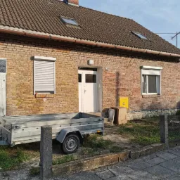 Parterowy dom z czerwonej cegły z białymi oknami i drzwiami, widoczne rolety zewnętrzne oraz przyczepka samochodowa z przodu.