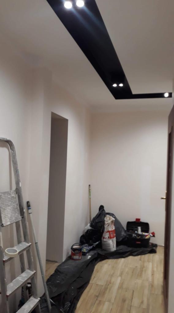 Wnętrze w trakcie prac w Czarnej: widoczny sufit z czarną wnęką oświetleniową, drabina malarska, podłoga zabezpieczona folią, narzędzia i materiały malarskie.