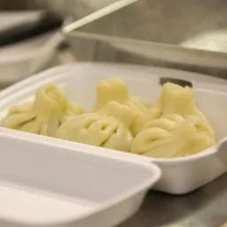 Chinkali w białym styropianowym pojemniku na wynos, widoczne fałdy ciasta i otwór na górze, gotowe do spożycia.