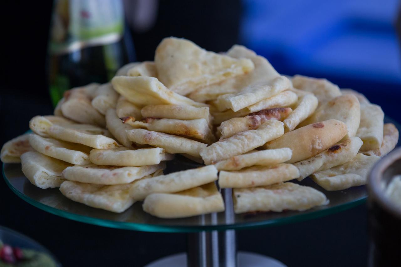 Stos jasnych, lekko przypieczonych placków na szklanym, okrągłym talerzu na metalowej nóżce, z butelką i miseczką w tle, sugerujący catering na imprezę.