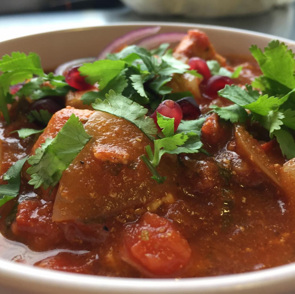 Gęsty gulasz warzywny z kawałkami mięsa, udekorowany świeżą kolendrą i pestkami granatu, podany w białej misce.