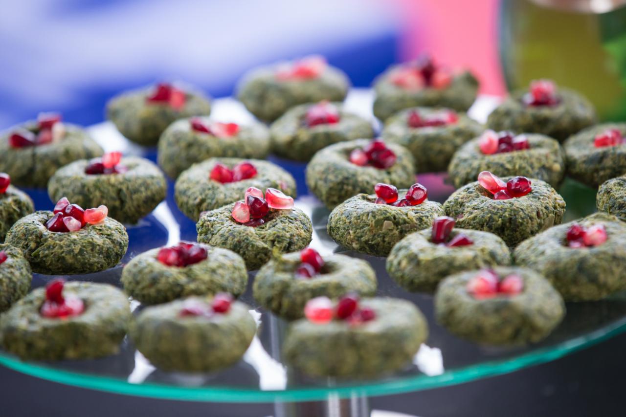 Szpinakowe kulebiaki udekorowane pestkami granatu, ułożone na szklanej paterze, widoczne w zbliżeniu, z rozmytym tłem w odcieniach niebieskiego i czerwonego.