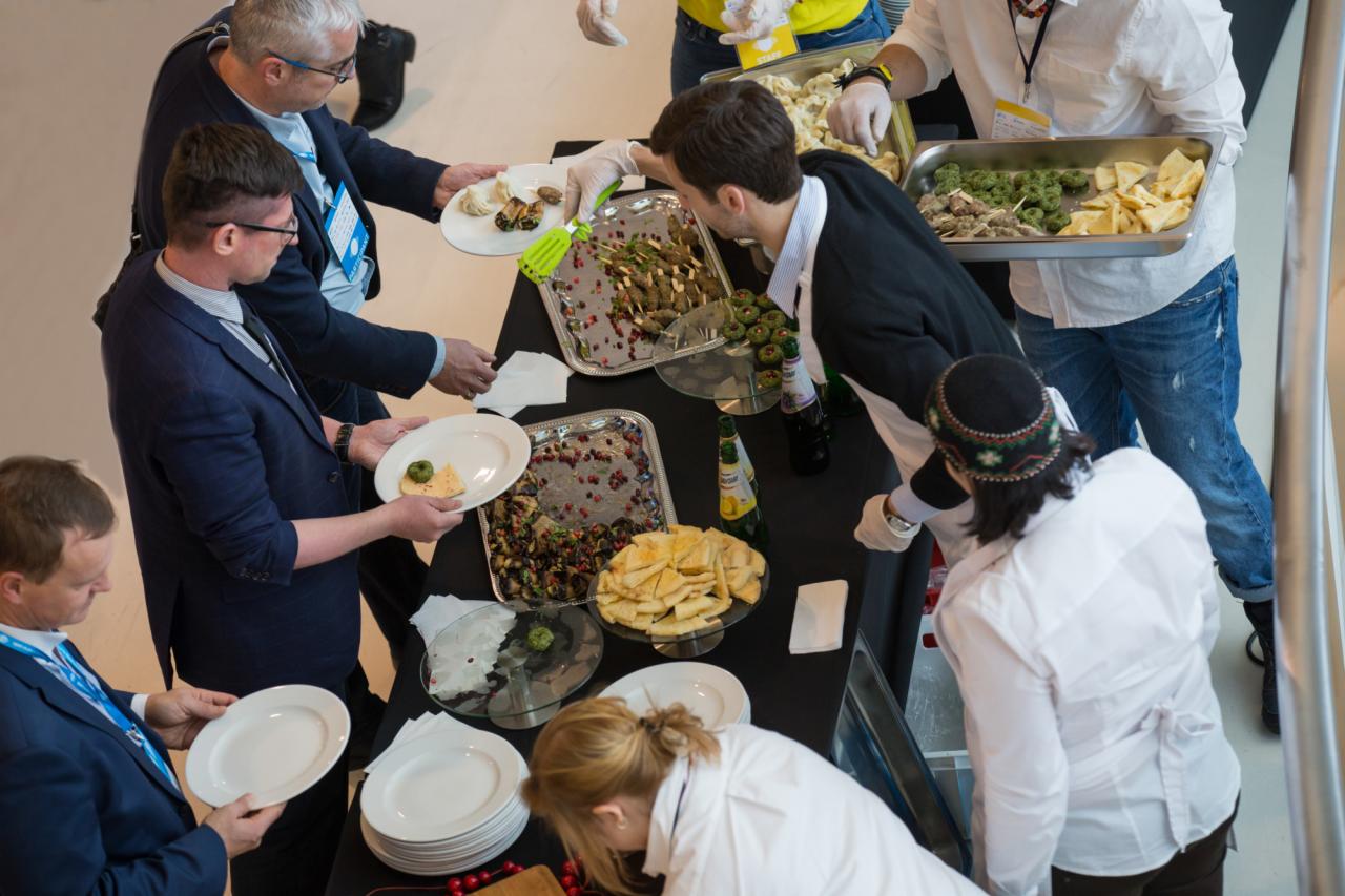 Widok z góry na stół z cateringiem: goście nakładają jedzenie z różnych tac, w tym szaszłyki, sałatki i przekąski serowe.