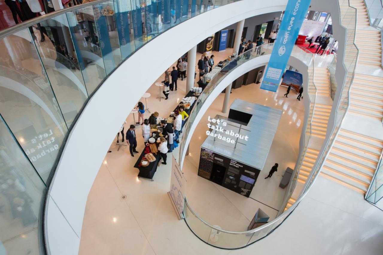 Widok z góry na atrium z cateringiem dla uczestników konferencji, z długim stołem bufetowym i napisem 'Let's talk about garbage'.