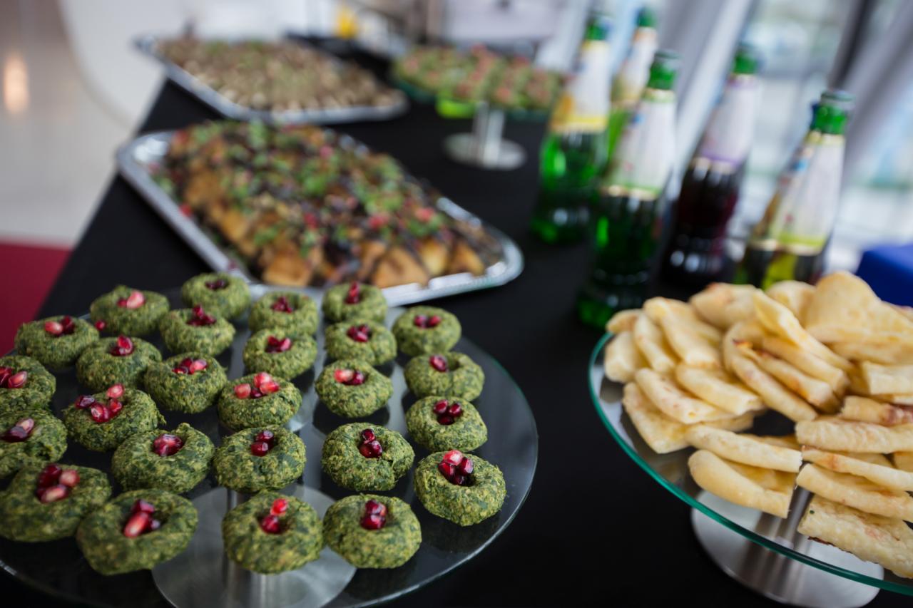 Stół z przekąskami: zielone kulki z granatem, placki, sałatki na srebrnych tacach i butelki z napojami w tle.