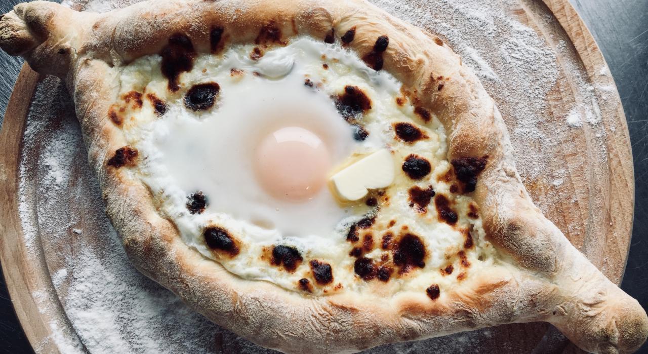 Adżarskie chaczapuri z jajkiem i masłem, podane na drewnianej desce posypanej mąką, z widocznymi przypaleniami sera i ciasta.