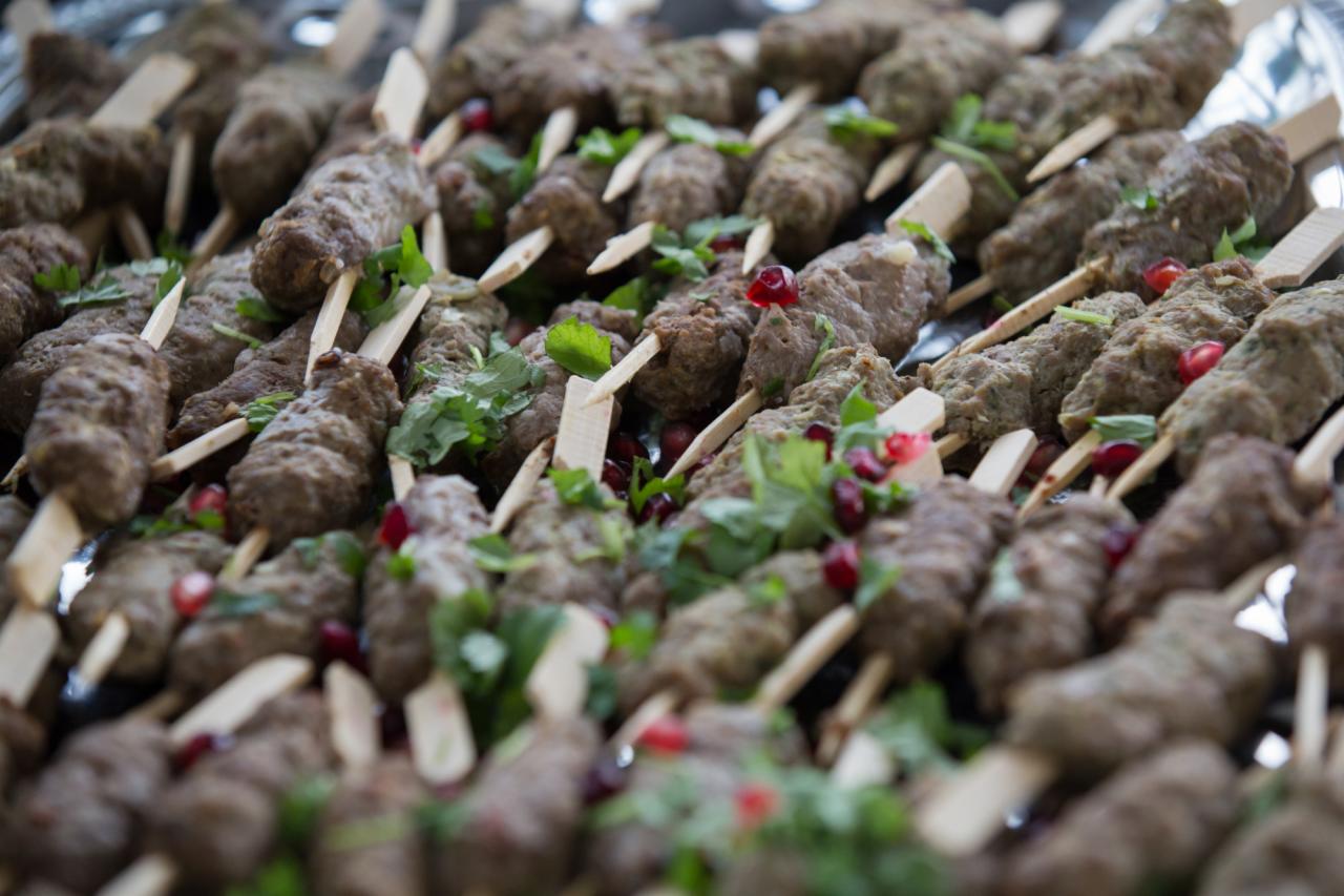 Szklana taca z rzędami mięsnych szaszłyków na drewnianych patykach, udekorowanych świeżymi ziołami i granatem.