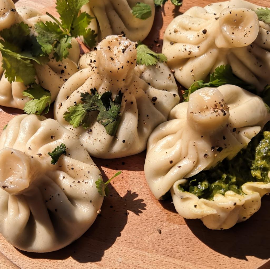 Gruzińskie pierożki chinkali posypane pieprzem i udekorowane świeżą kolendrą, jeden rozcięty ukazujący zielone nadzienie, ułożone na drewnianej desce w ciepłym świetle.