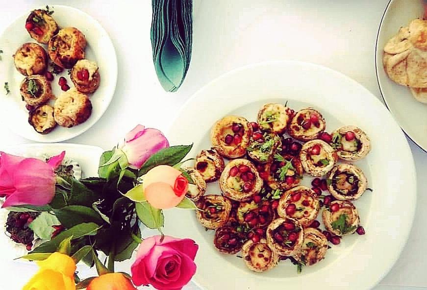 Elegancko podane grzyby nadziewane farszem i posypane granatem na białym talerzu, obok bukiet róż w odcieniach różu i żółci, oraz złożone serwetki w kolorze zielonym. Ujęcie z góry.