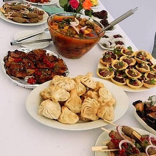 Stół z różnorodnymi daniami: zupa w szklanej misie z łyżką, pierożki chinkali, szaszłyki na patyku z czerwoną cebulą i granatem, mięso z granatem, tartaletki z mięsem i ziołami, grillowane warzywa.